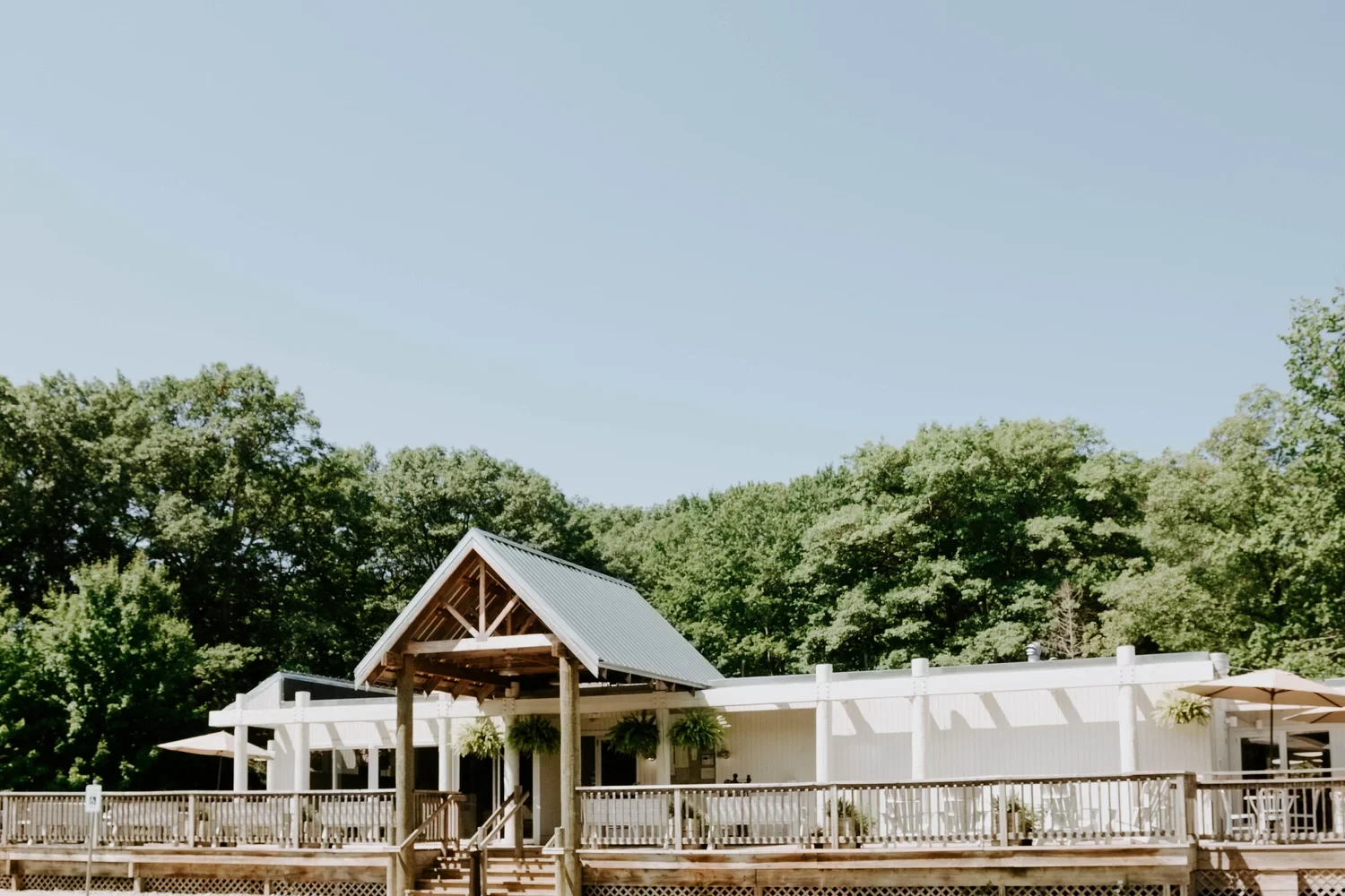 Wide view of Sandy Point Beach House’s white building, peaked entrance and front deck beside the trees